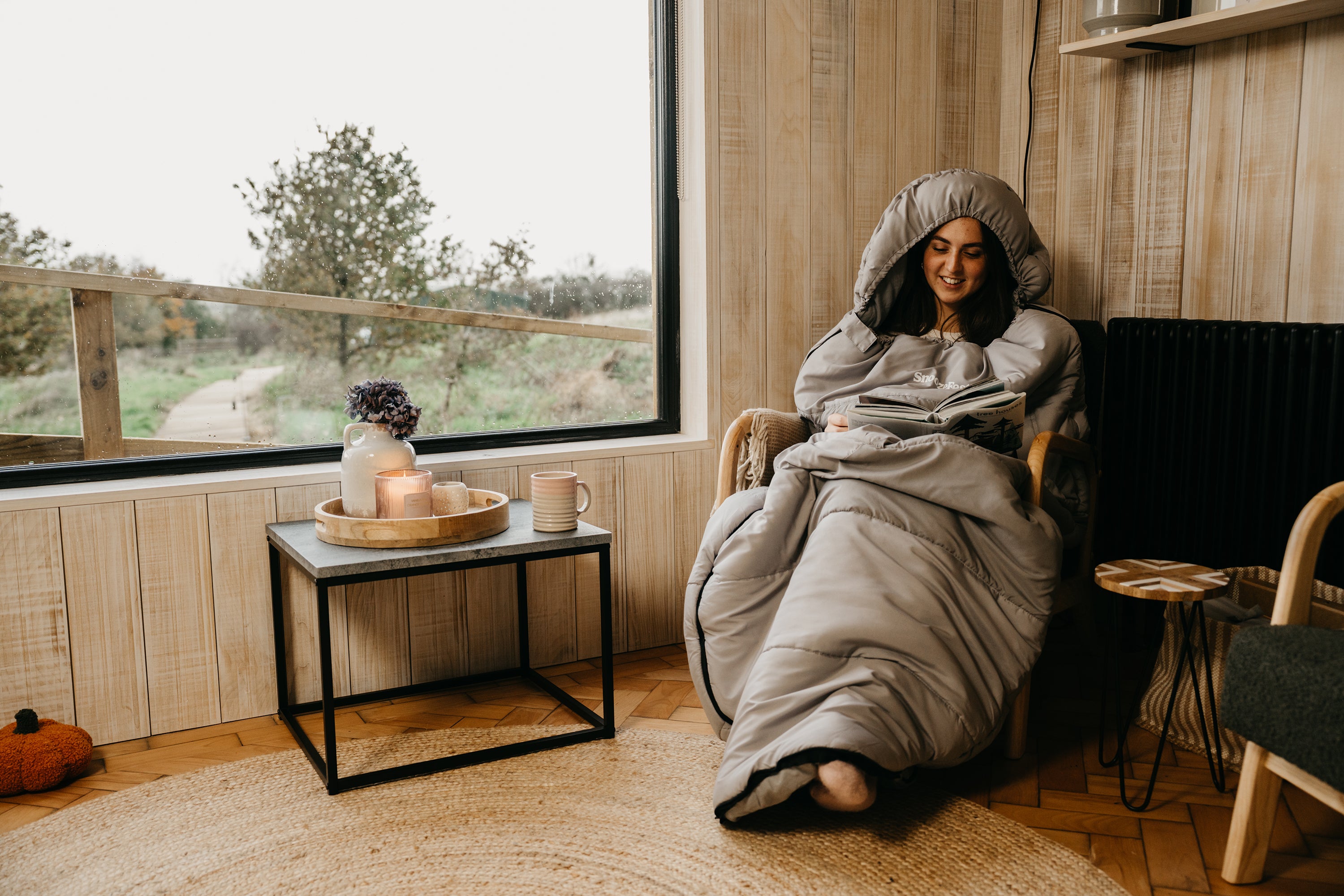 Person in a wearable blanket sitting in a cozy room with a window view.