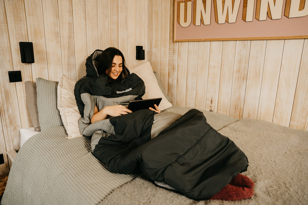Person lying on a bed using a tablet with a wearable blanket on them, in a room with wooden walls and 'UNWIND' sign.