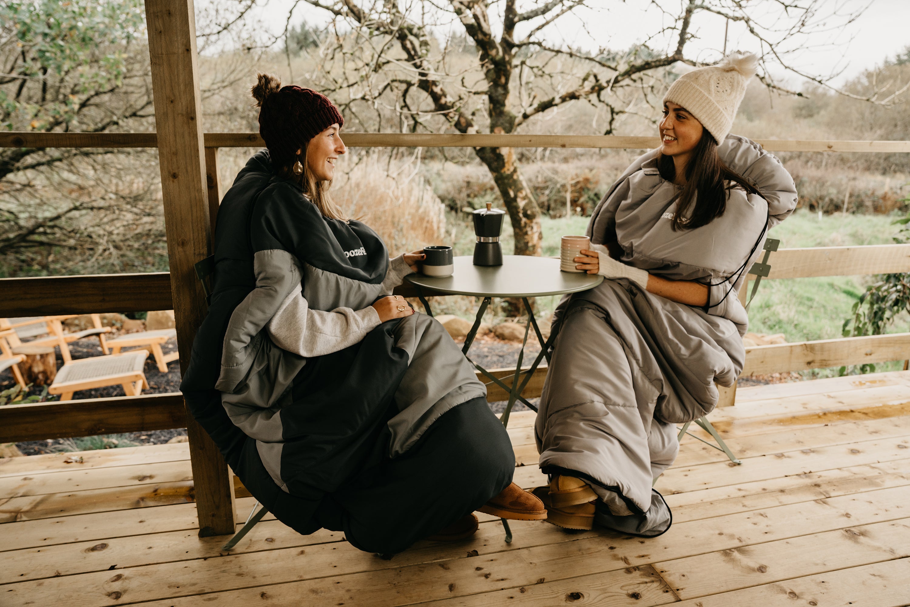Two people sitting on a wooden deck, wrapped in wearable blankets, enjoying coffee.