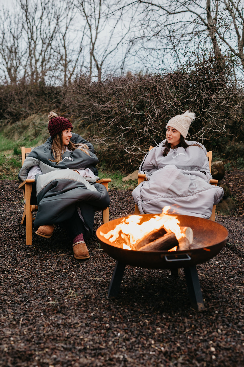 Two people in wearable duvets, sitting by a fire pit in a natural setting.