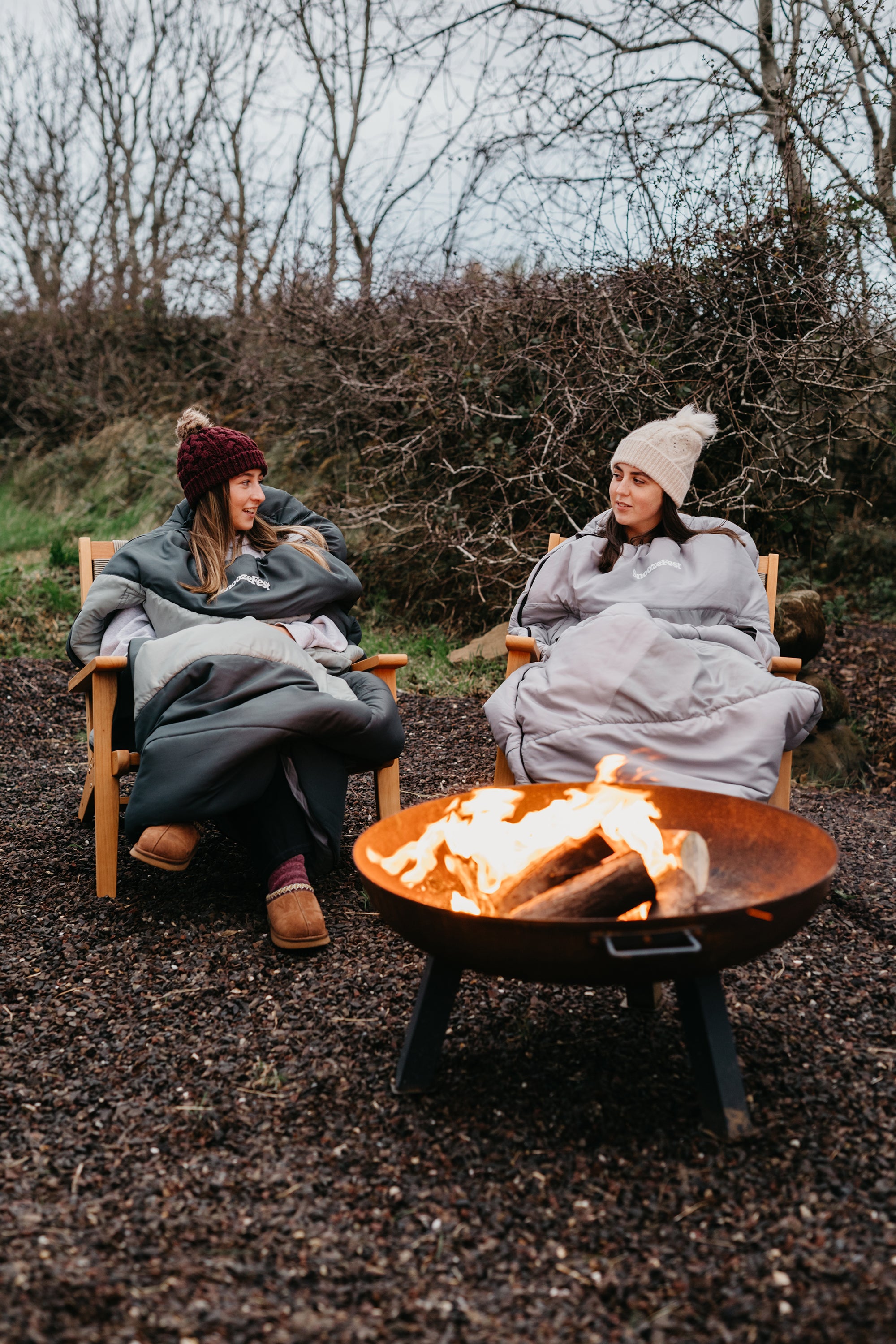 Two people in wearable duvets, sitting by a fire pit in a natural setting.
