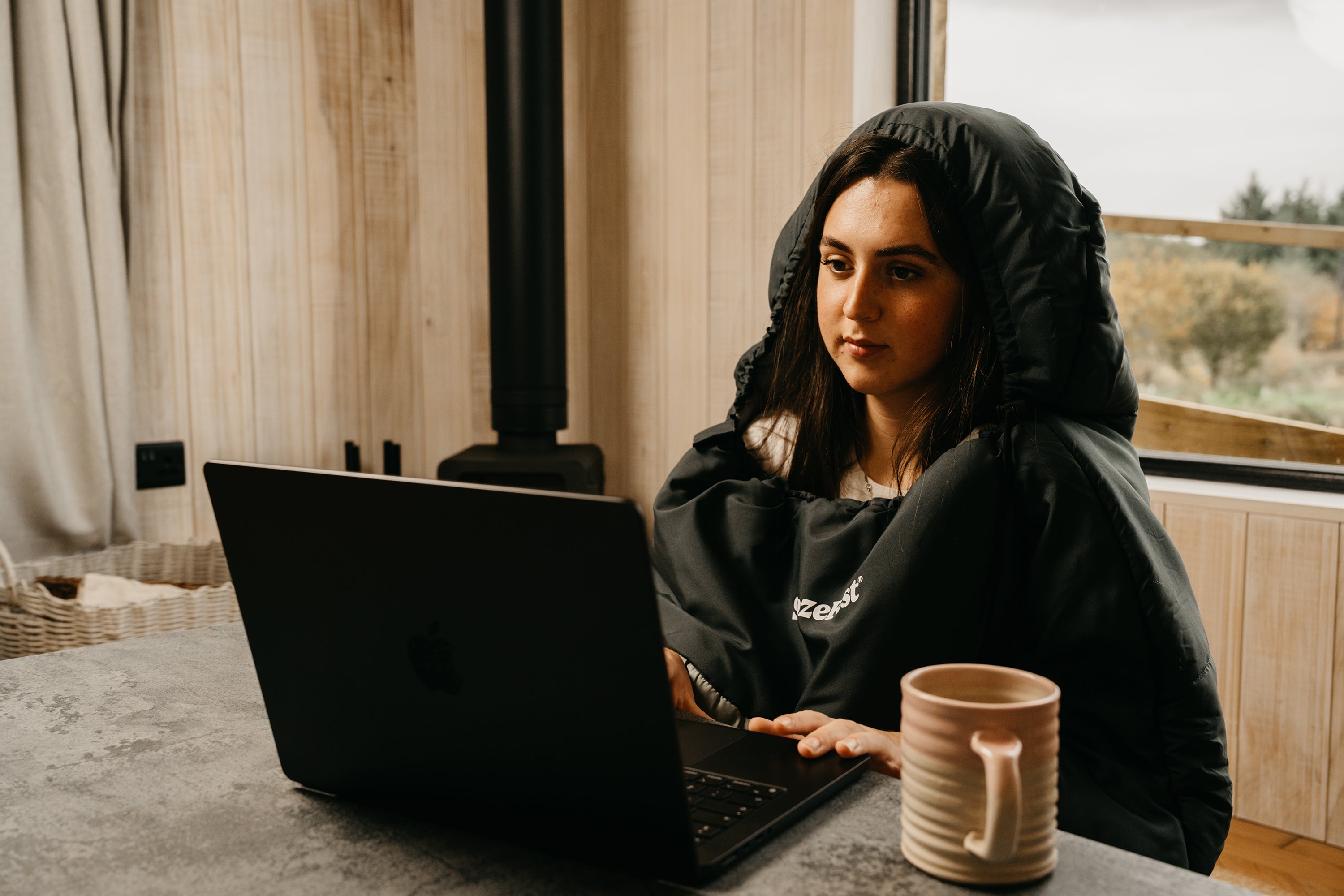 Person sitting at a table with a laptop and a mug, in a wearable blanket