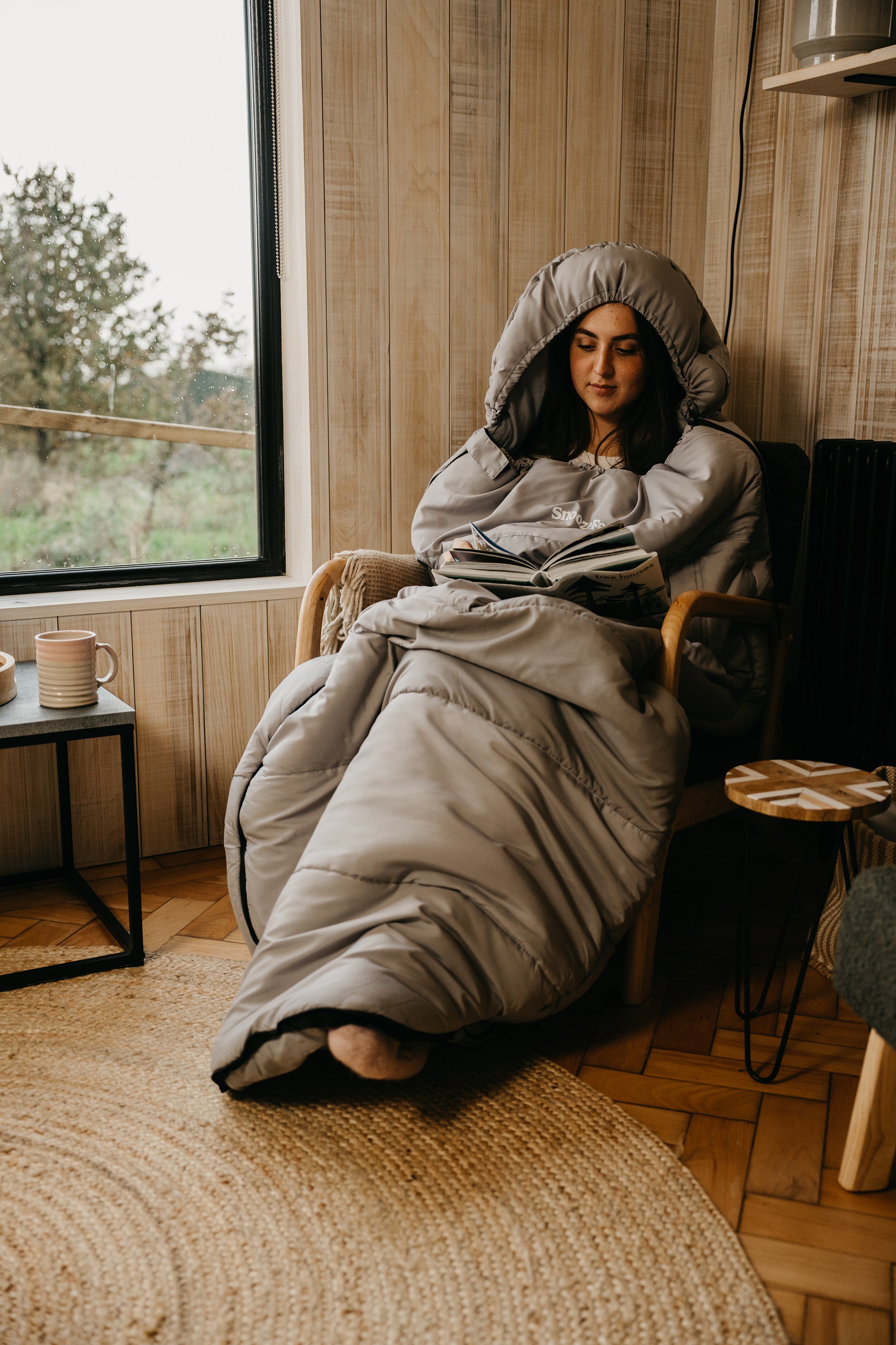 Person wrapped in a wearable gray hooded duvet sitting on a chair by a window.