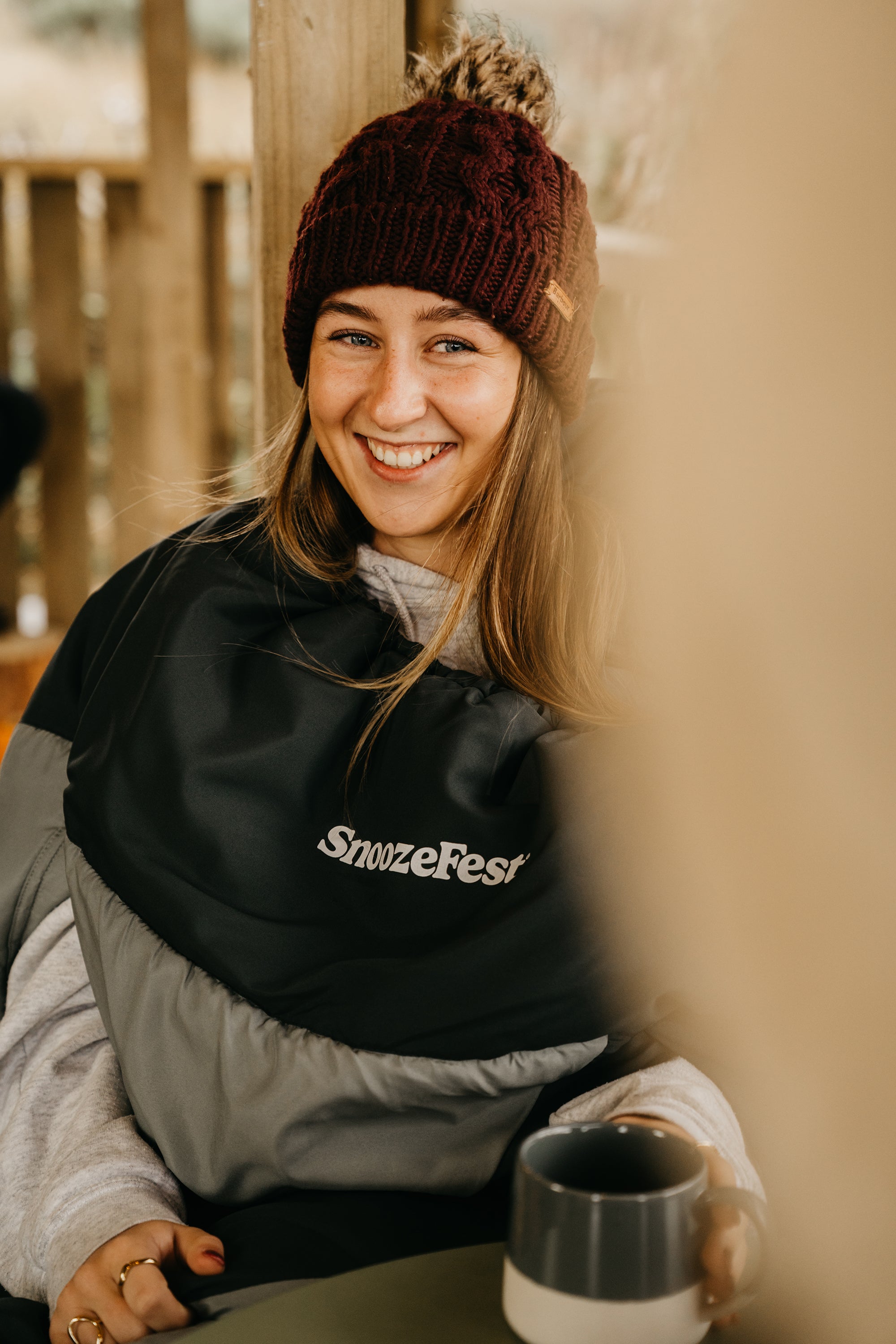 Person in a wearable duvet with 'SnoozeFest' logo, holding a mug outdoors.