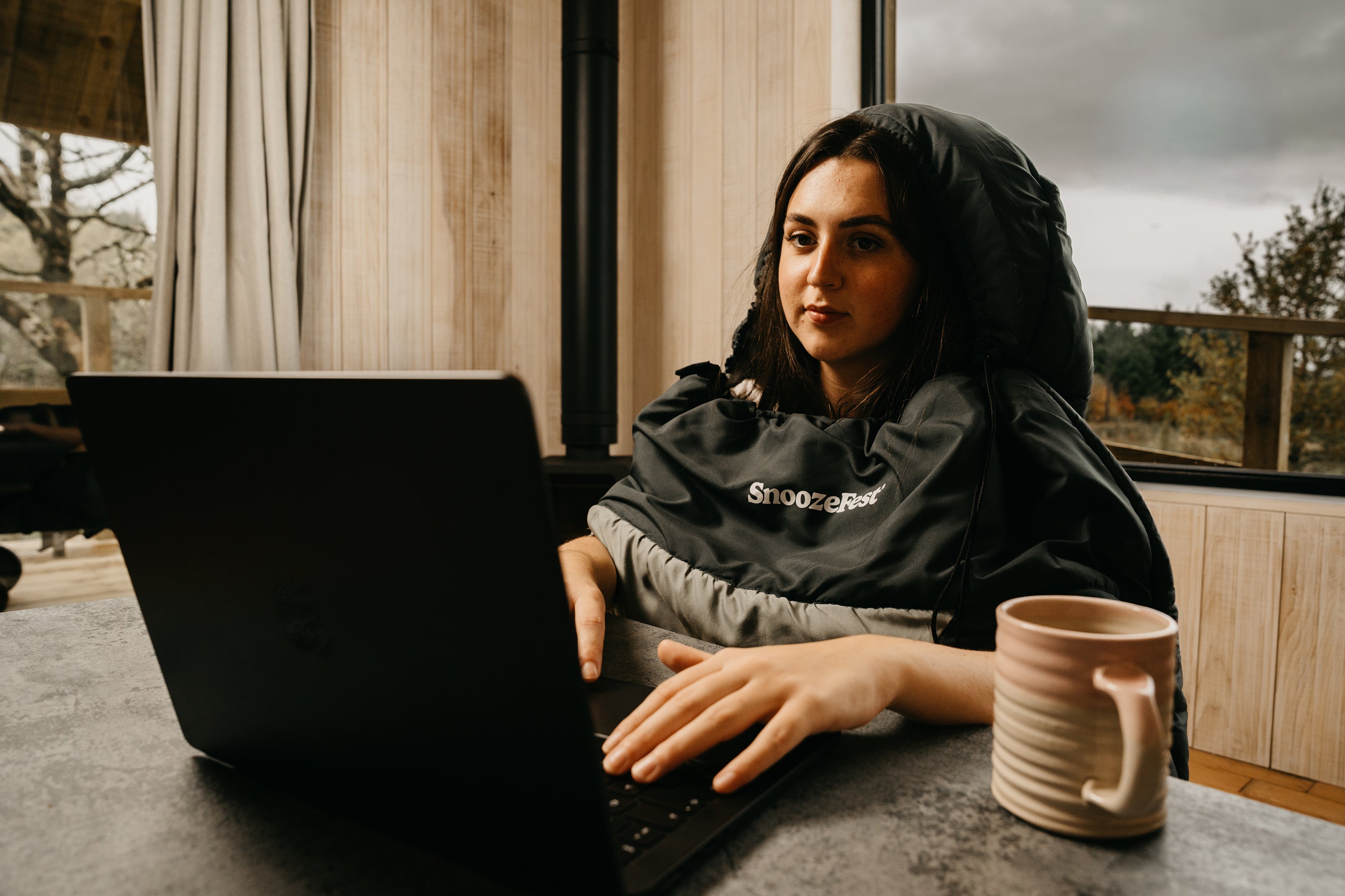 Person working from home, at a table with a laptop and a mug, in a cozy wearable blanket.