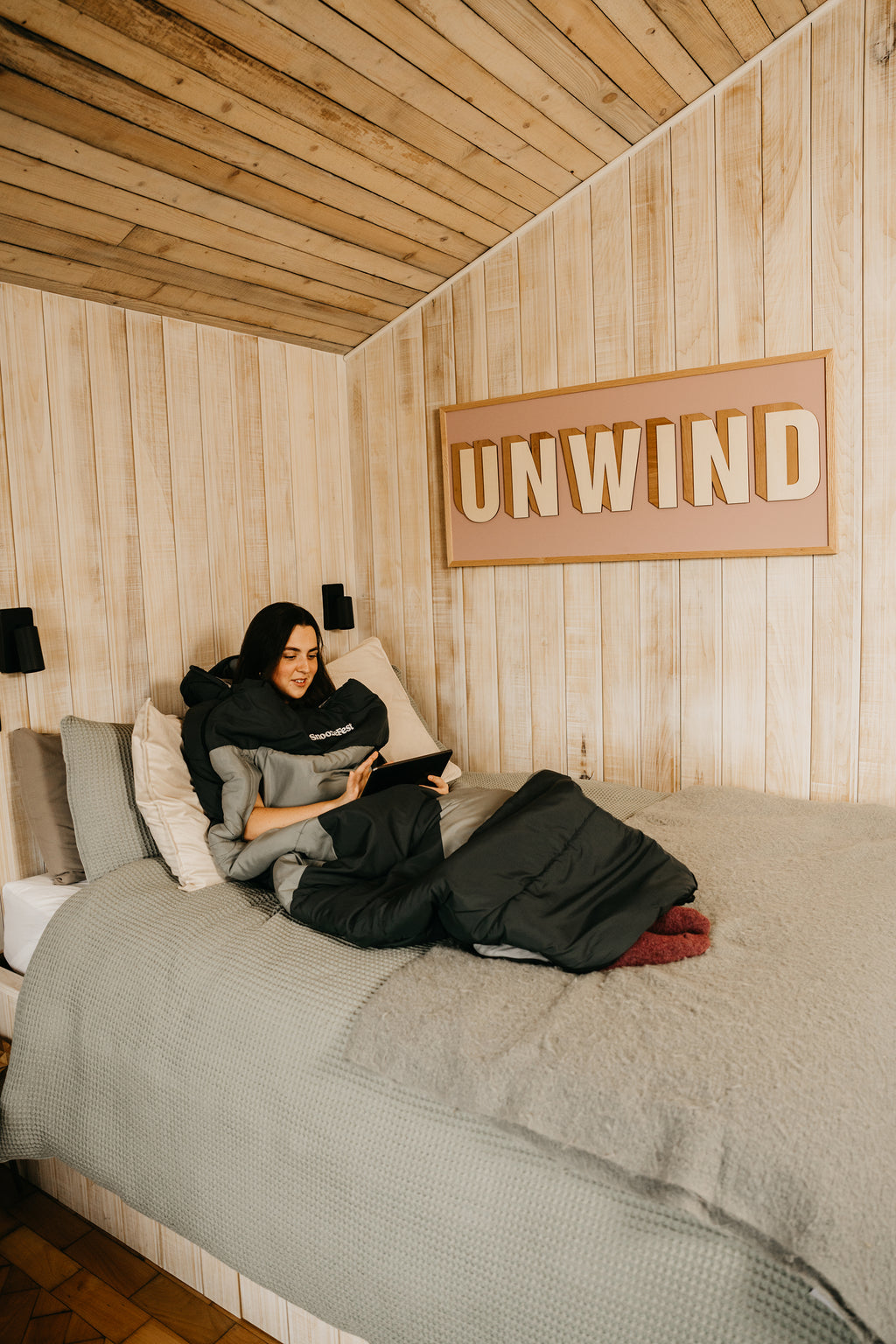Person in a wearable duvet, sitting on a bed in a wooden cabin with 'UNWIND' sign above.