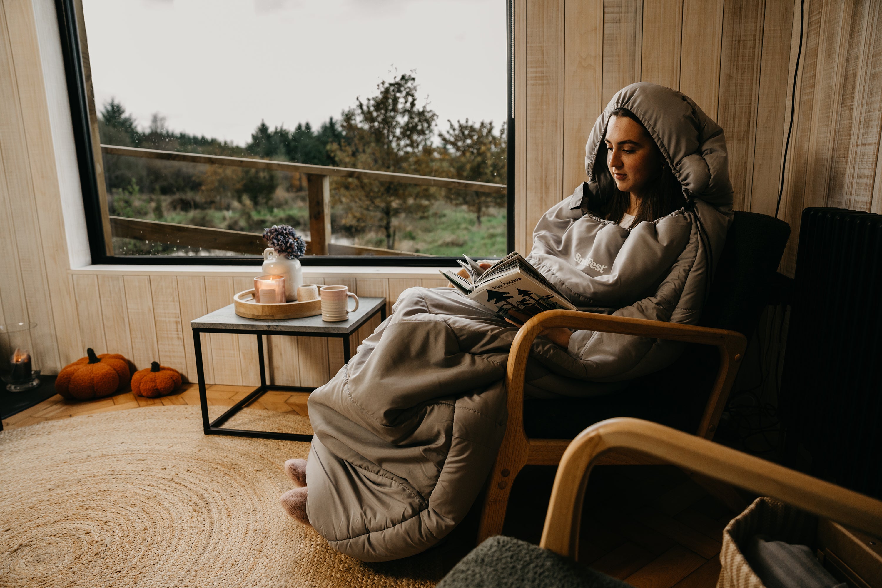 Person wearing a wearable blanket, sitting in a cozy room, surrounded by autumn decorations.