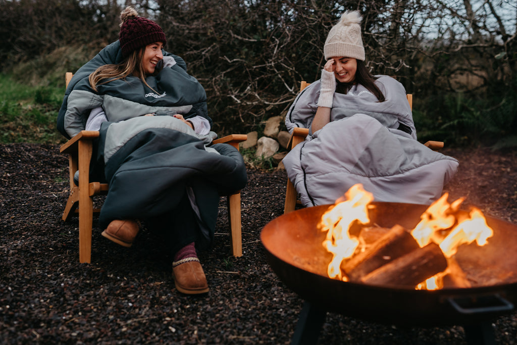 Two people sitting by a fire pit, wrapped in wearable duvets in a natural setting.