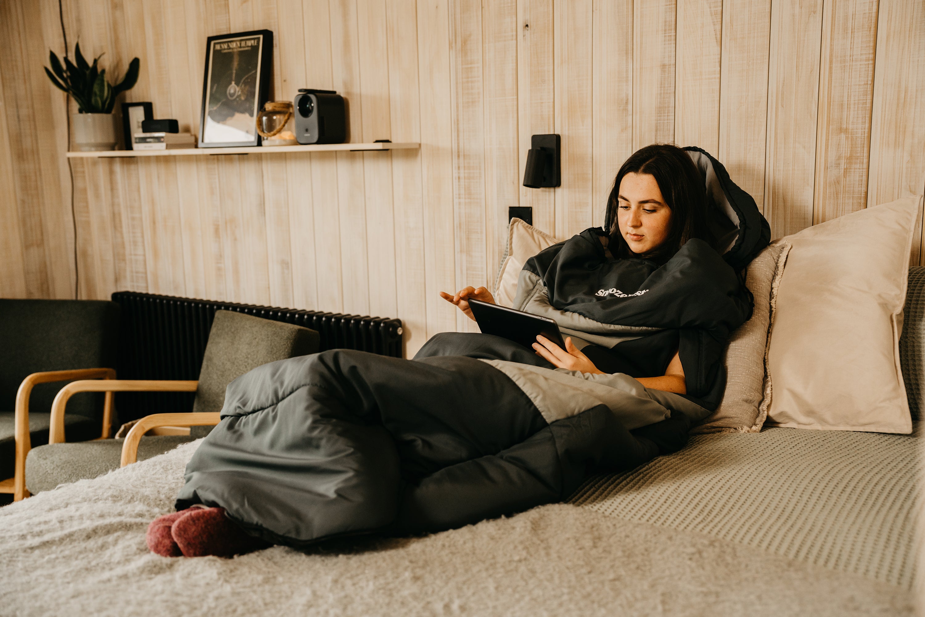 Woman wearing a SnoozeFest pod, lying on a bed using a tablet in a cozy room with wooden walls and a shelf.