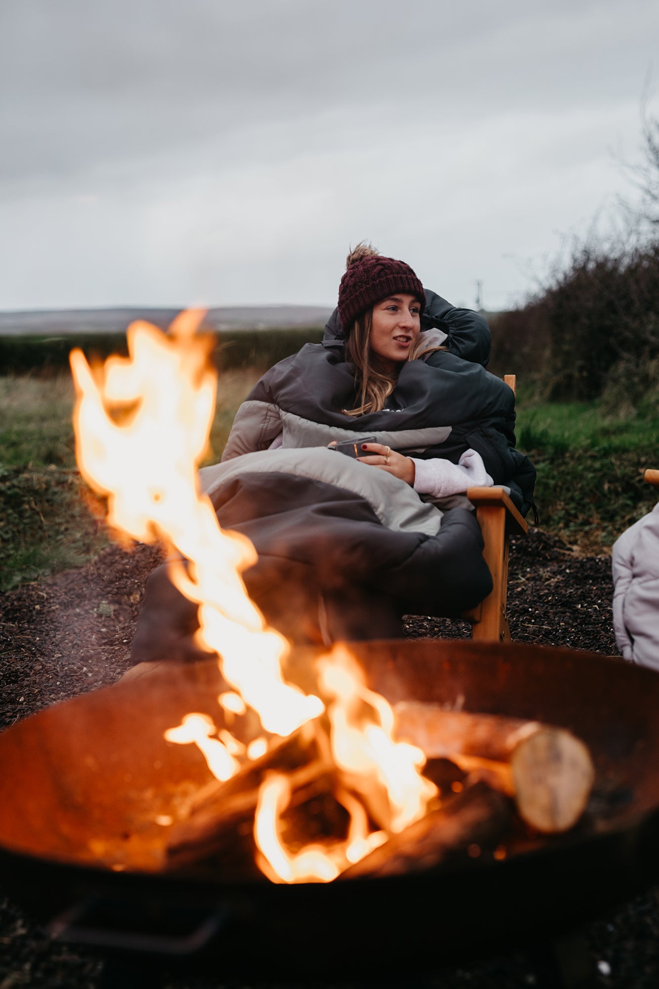 Young person, staying cosy outdoors, sitting by a campfire, wearing a SnoozePod, from SnoozeFest
